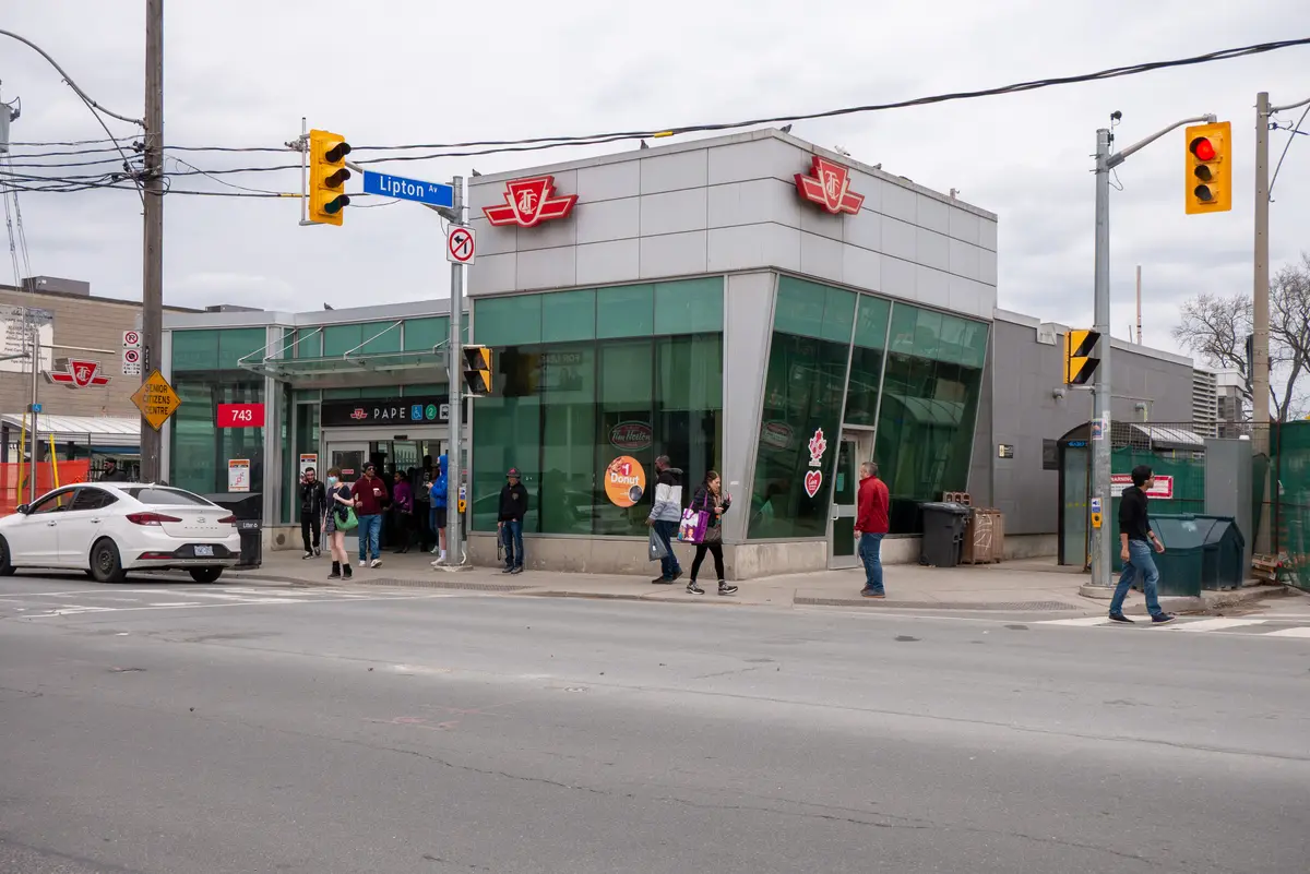 The glass and steel entrance of Pape subway station on Pape Avenue, with commuters walking past
