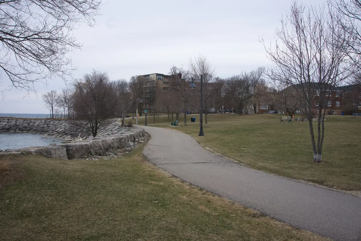 A paved path curving along the lakeshore at Mimico Waterfront Park, with a rock breakwater, bare trees and apartment buildings behind