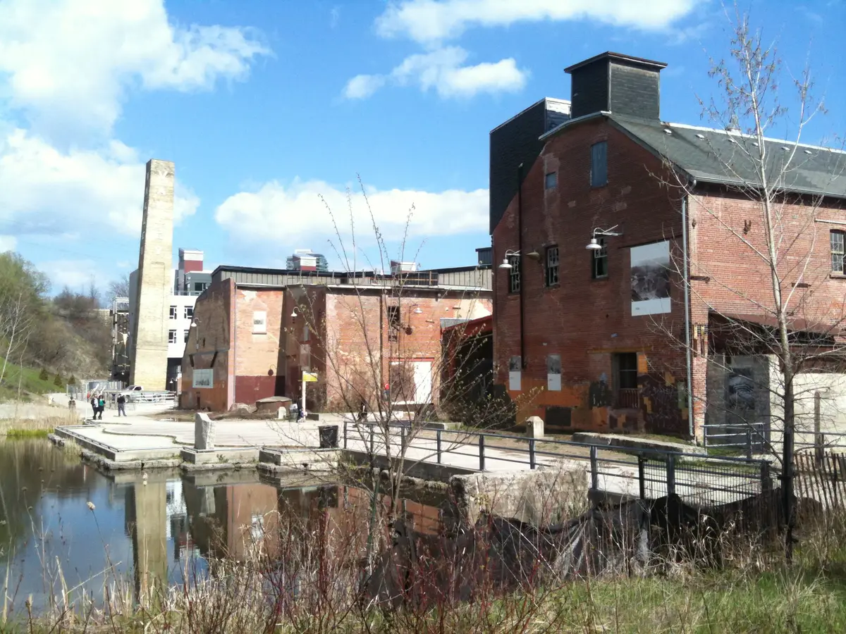 Red brick factory buildings with a tall chimney beside a pond and a footbridge, under blue sky with bare trees
