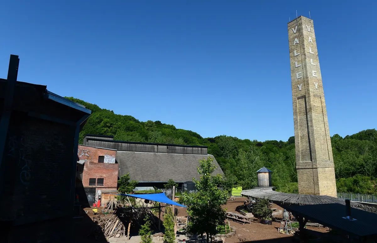 A tall brick chimney with vertical lettering rises above the brick sheds and yard of Don Valley Brick Works, with a wooded hillside behind