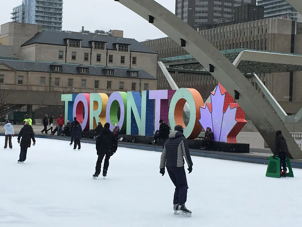 Skaters on an outdoor rink in front of a large, multicoloured TORONTO sign.