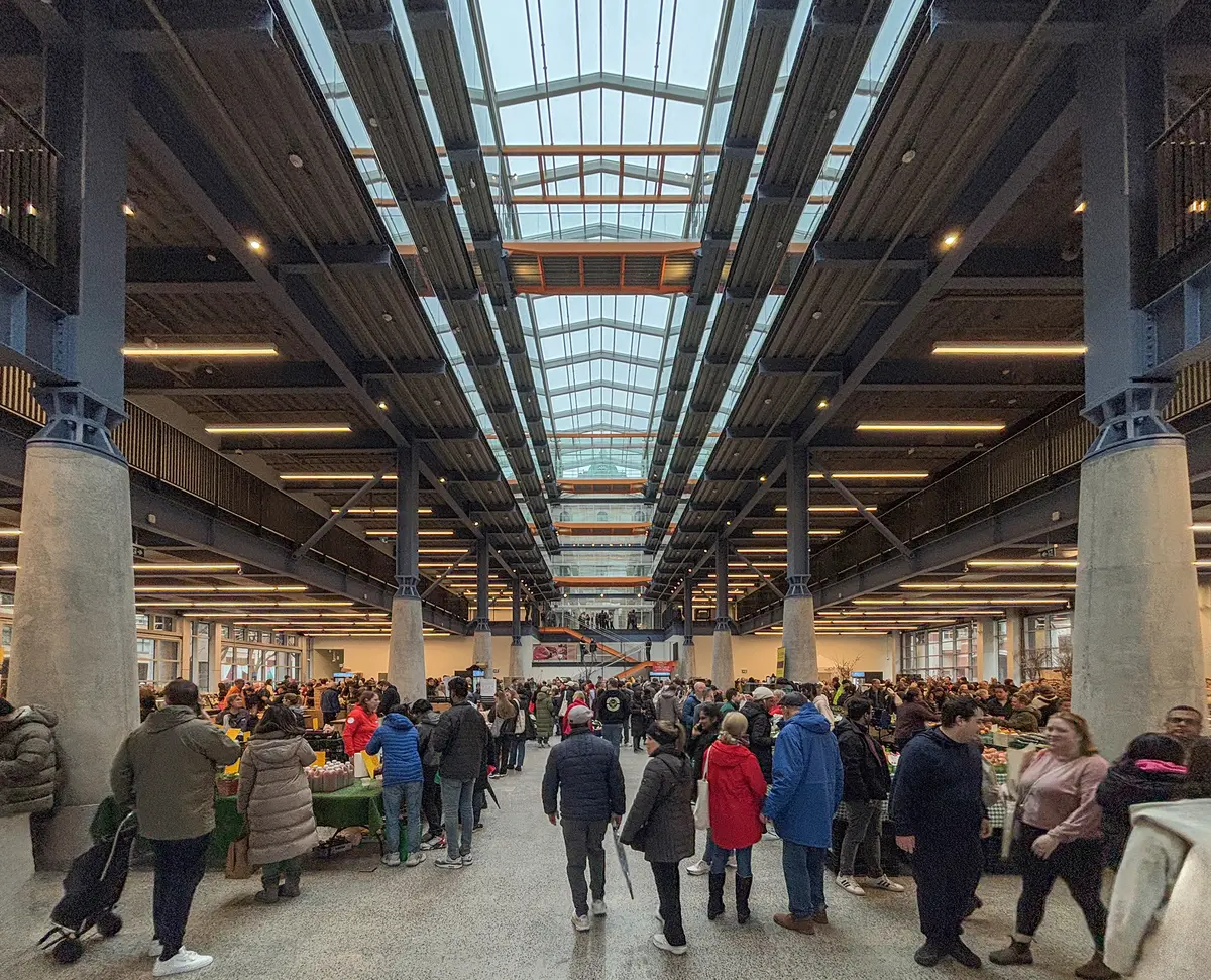 Shoppers move between market stalls under the glass roof and steel trusses of St. Lawrence Market North.