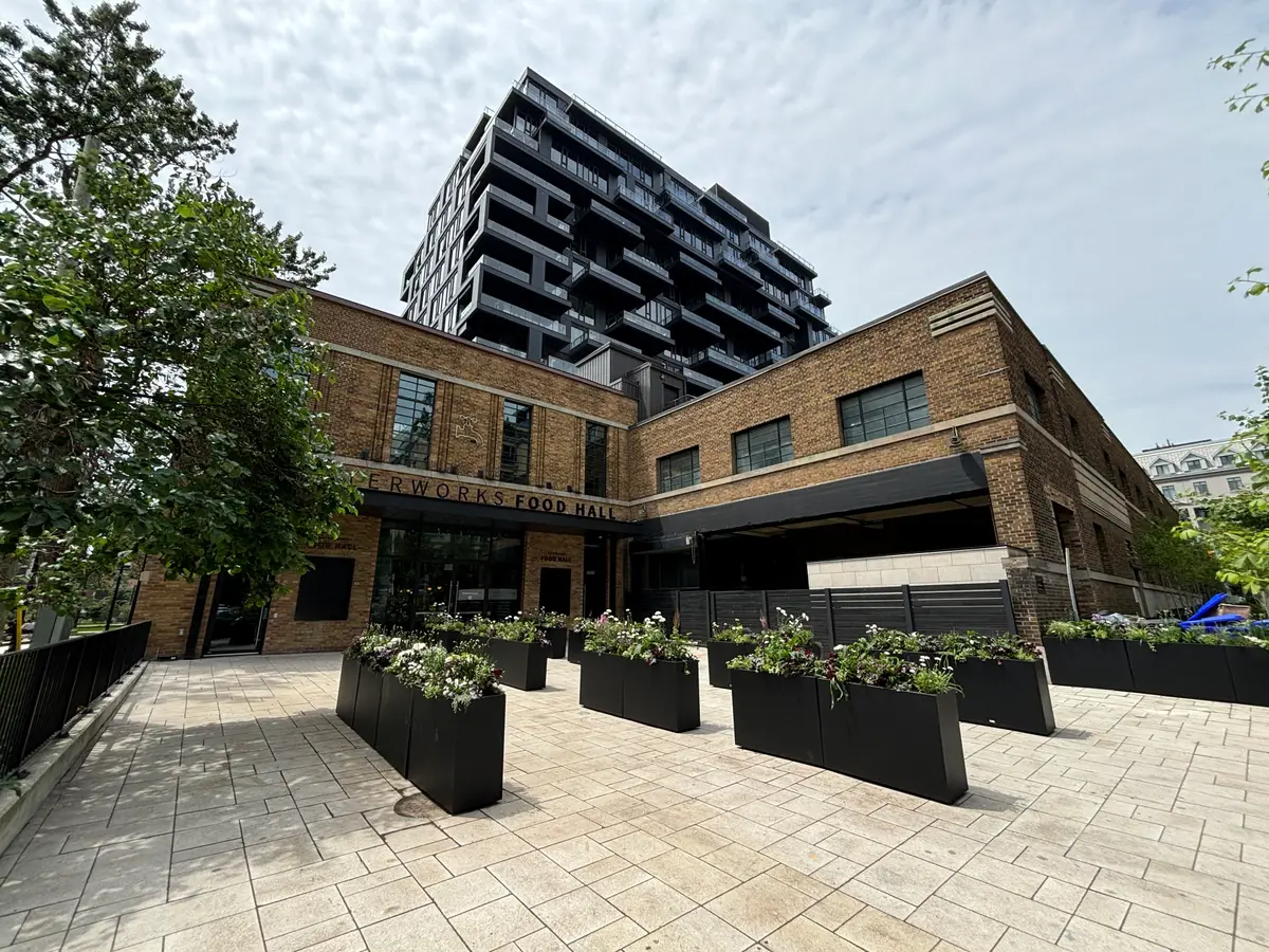 A paved courtyard with planters leading to the brick entrance of Waterworks Food Hall, with a condo tower above