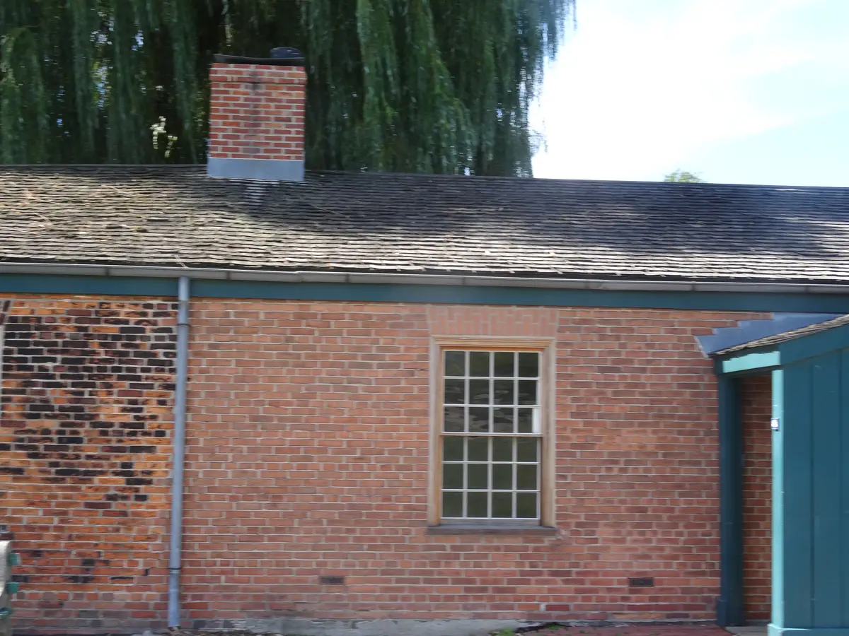 A brick building with a multi-pane window and a shingled roof, with a weeping willow behind it