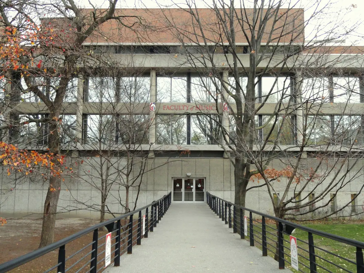 A concrete and glass university building with Faculty of Music lettering, reached by a walkway among autumn trees