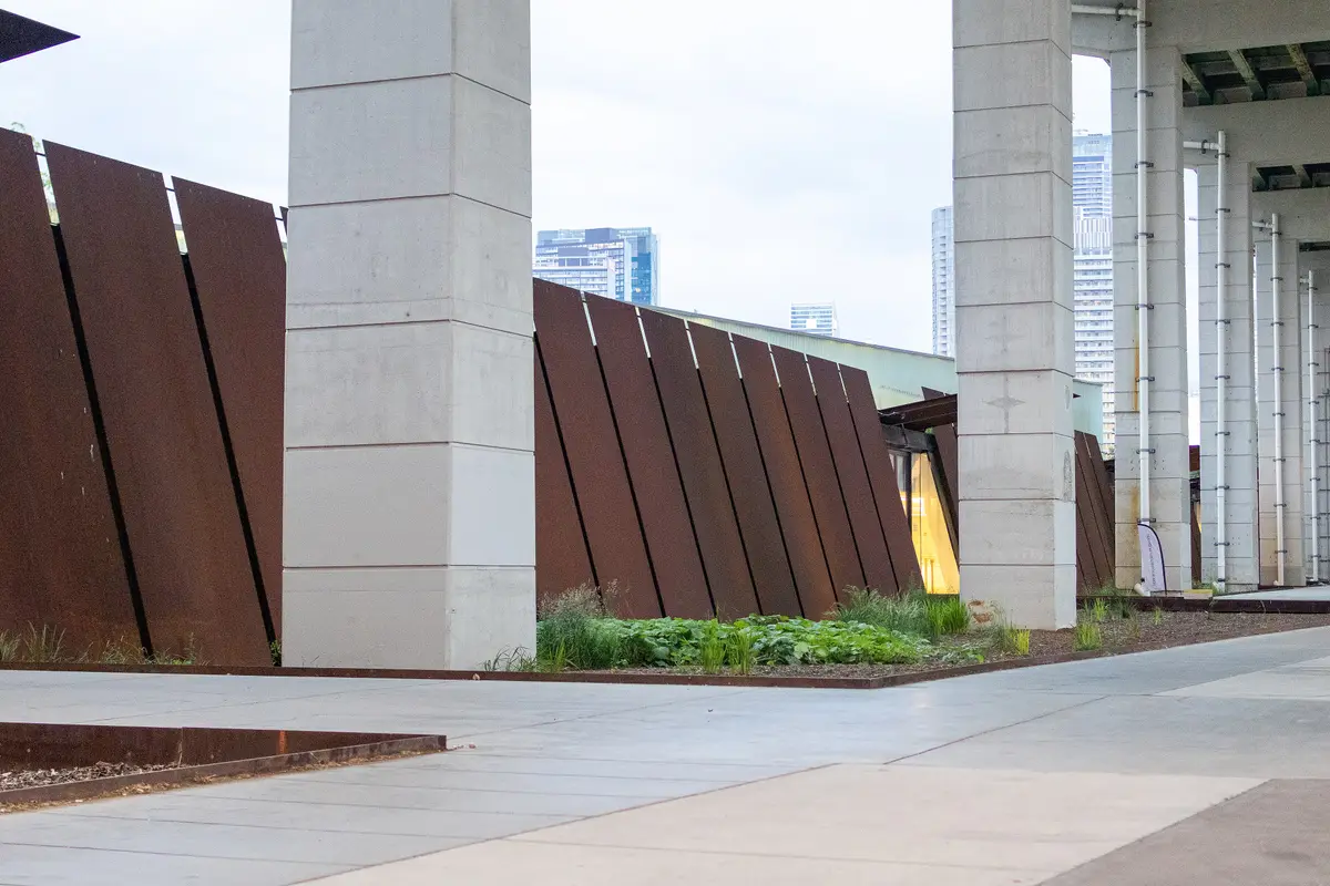 The weathered steel wall of the Fort York visitor centre beside tall concrete pillars, with condo towers behind.