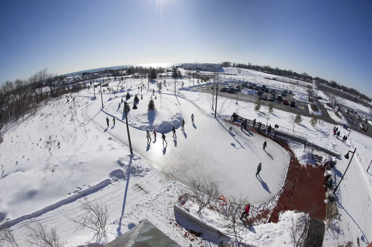 A curving outdoor skating trail seen from above, with skaters on the ice and snowy parkland around it