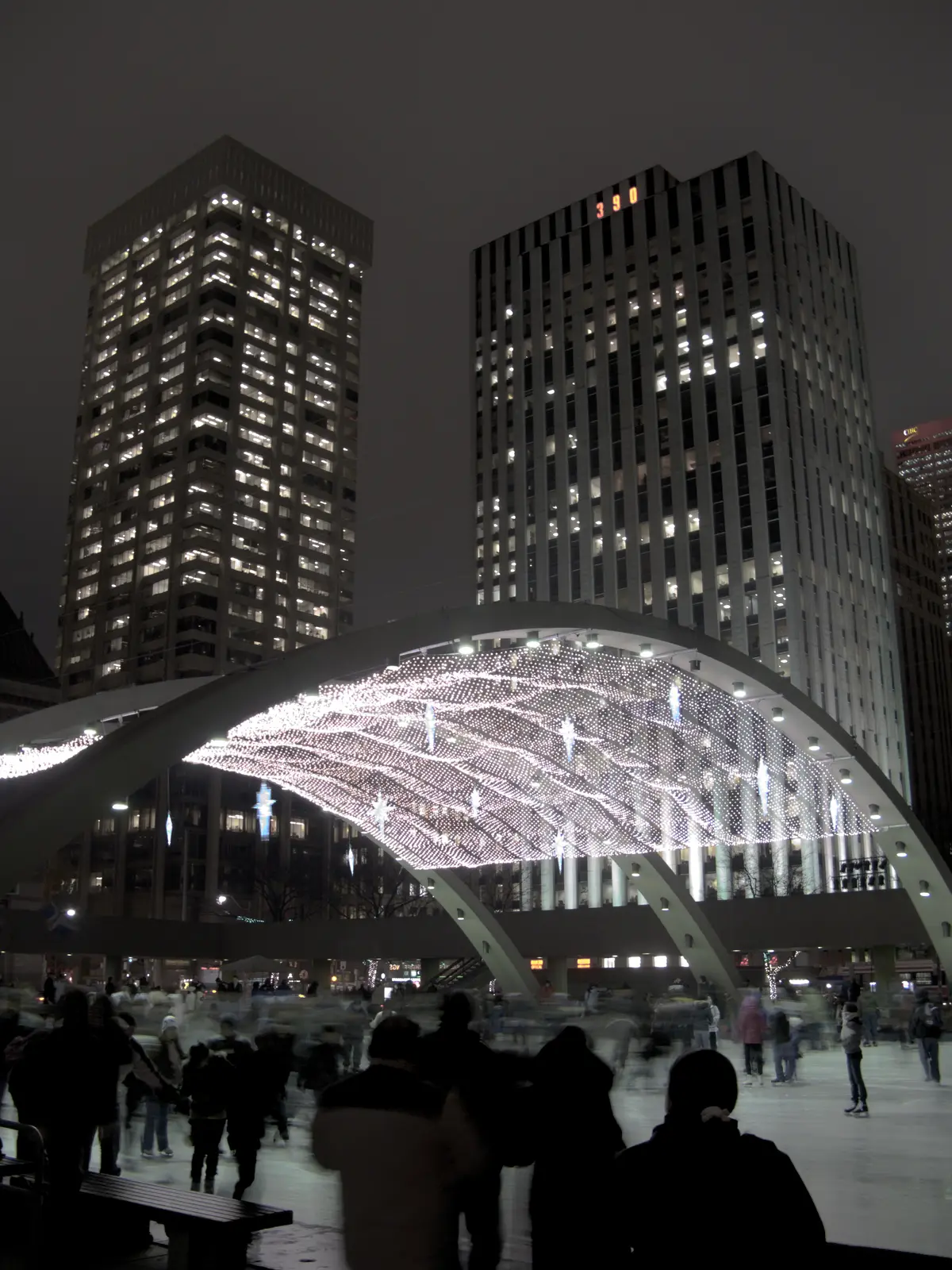 Skaters blurred in motion on a night rink beneath a lit arch, with office towers behind