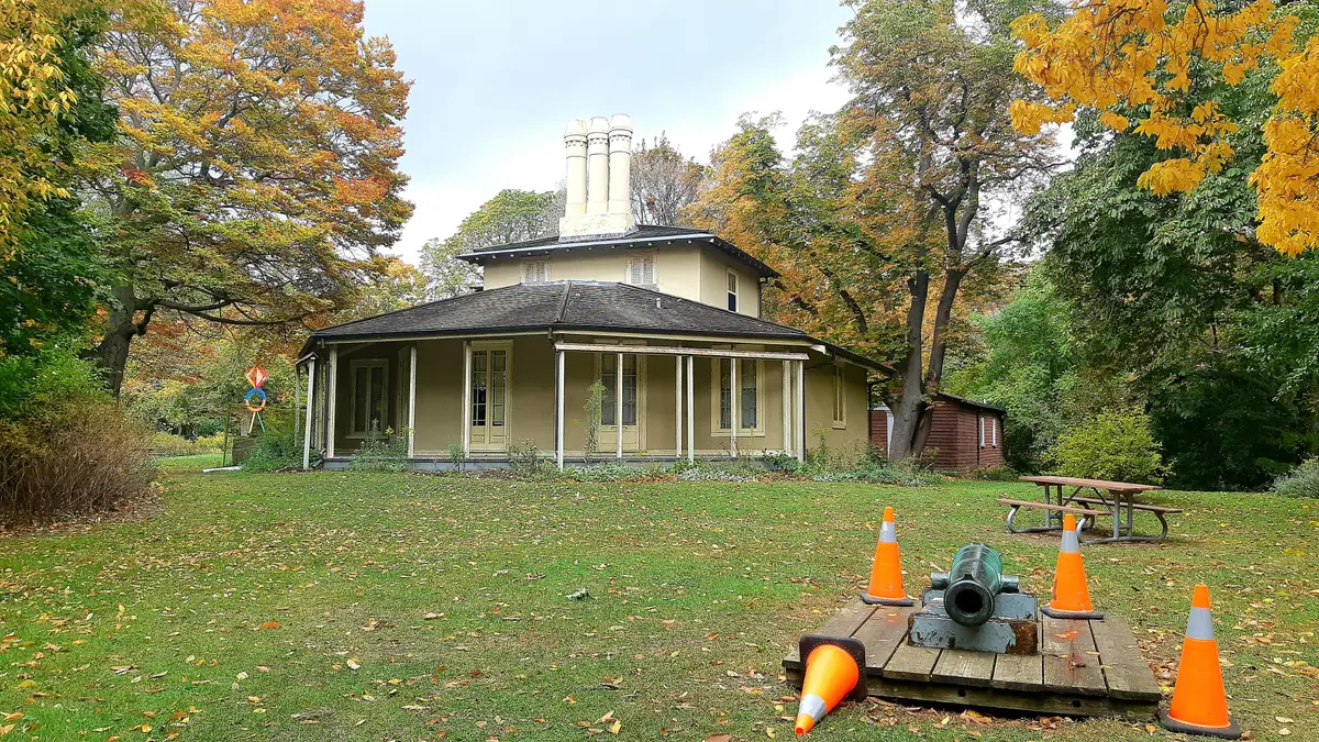 Colborne Lodge in High Park, a low cream house with a veranda and three white chimneys, surrounded by autumn trees and a cannon on a pallet.