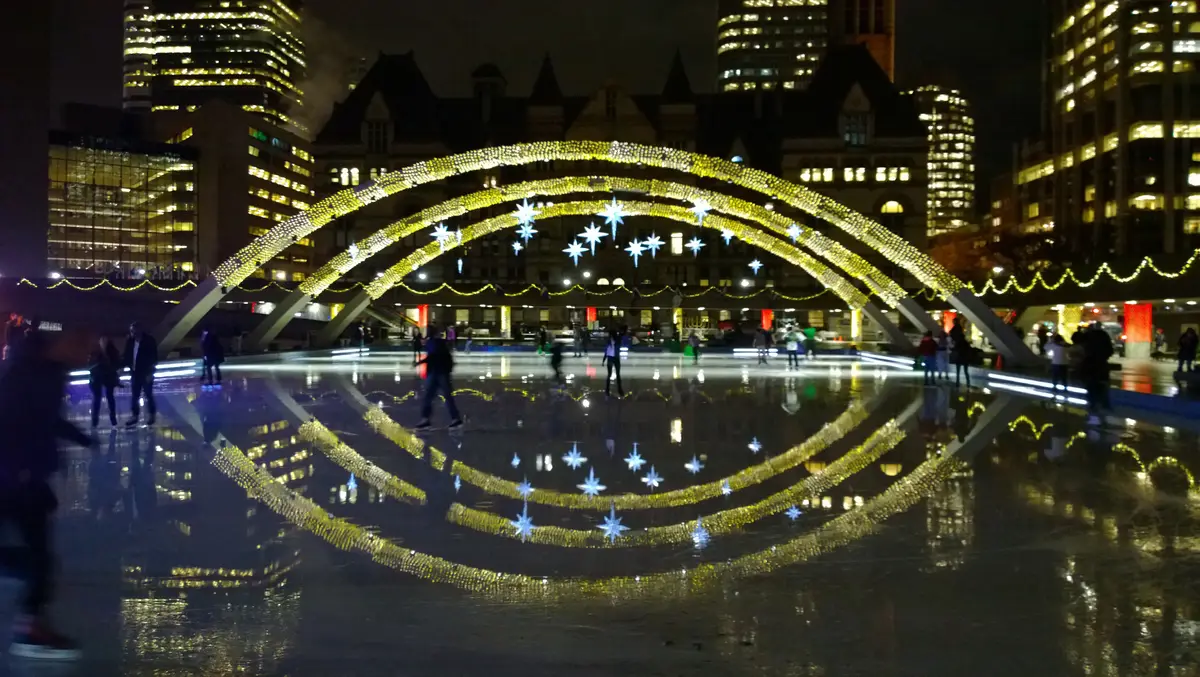 The rink at Nathan Phillips Square at night, with the lit arches reflected in the ice and skaters gliding beneath City Hall.