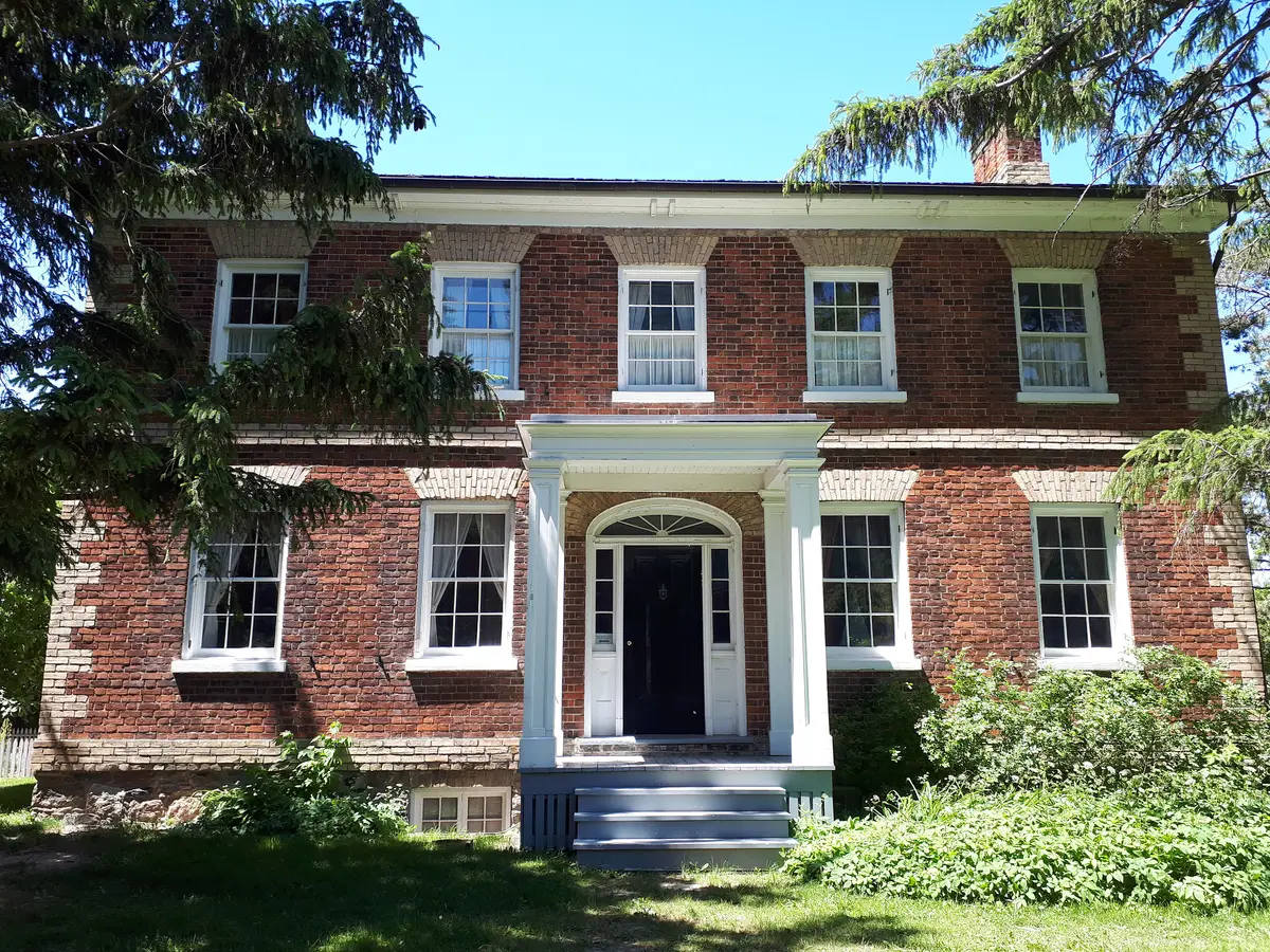 The red brick front of Gibson House with a white porch, tall windows and evergreen trees