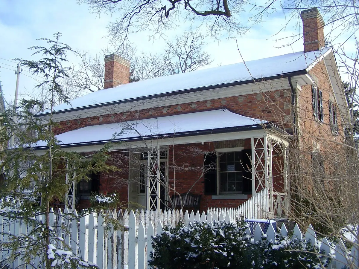 A brick farmhouse with snow-covered roofs and a white picket fence among bare trees