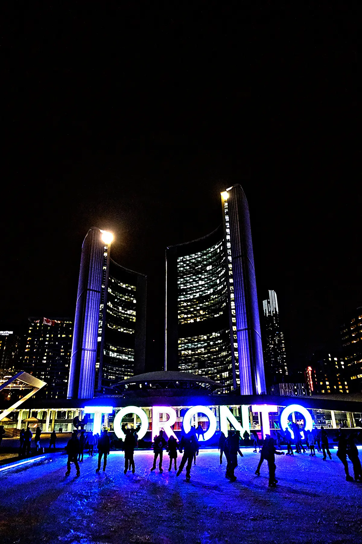 Skaters on blue-lit ice in front of the glowing TORONTO sign and the curved towers of City Hall at night