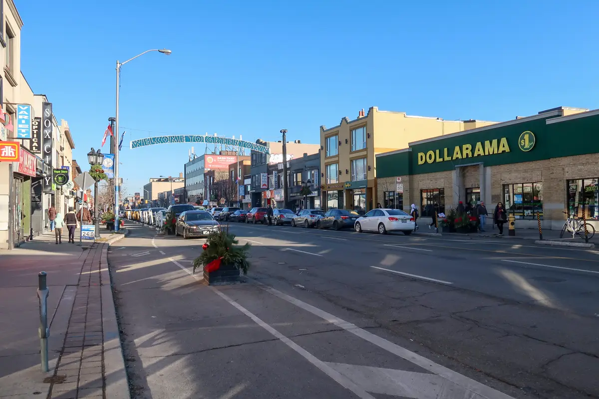 A Welcome to Greektown sign arching over Danforth Avenue under a clear blue sky, with shopfronts and parked cars along the street
