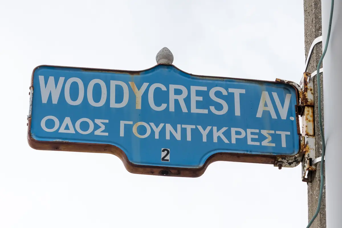 A blue street sign reading Woodycrest Avenue with the street name repeated in Greek letters below