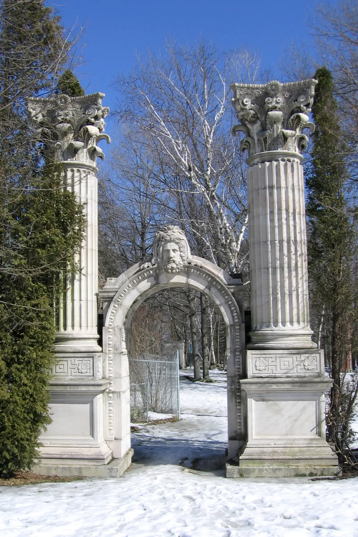Two tall stone pillars with carved capitals flanking an arch, with snow on the ground and bare birches behind.
