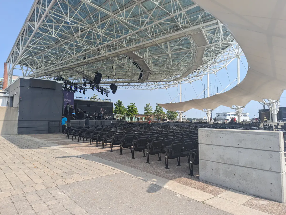 A covered outdoor stage with steel roof trusses and rows of empty seats on a sunny day