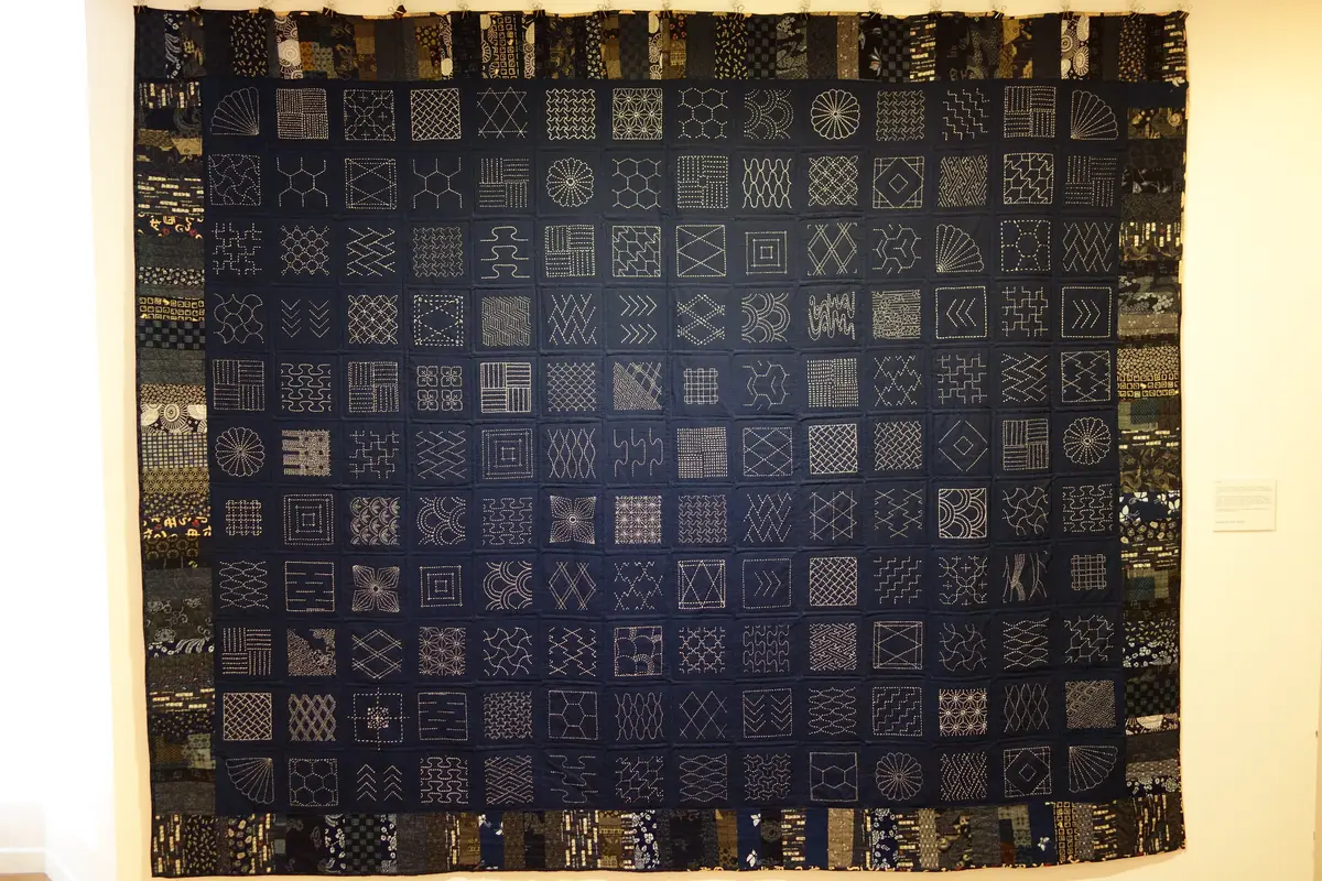 A large dark indigo quilt stitched with rows of white geometric sashiko patterns, hung on a gallery wall