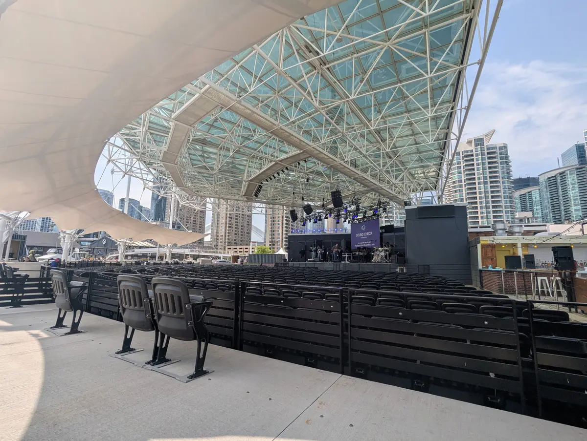 A covered outdoor stage with a steel-framed roof and rows of seating at Harbourfront Centre, with residential towers beyond