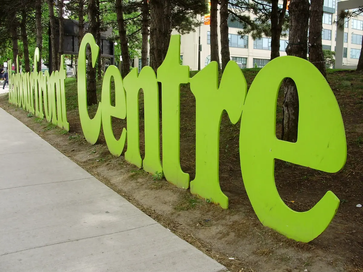 Large lime-green Harbourfront Centre letters on a strip of lawn beside a sidewalk and trees.