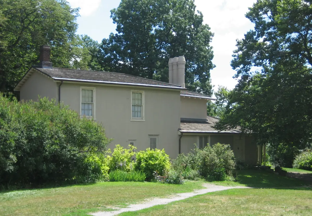 Two-storey pale cream cottage with tall chimneys and a gravel path across the lawn, framed by mature trees