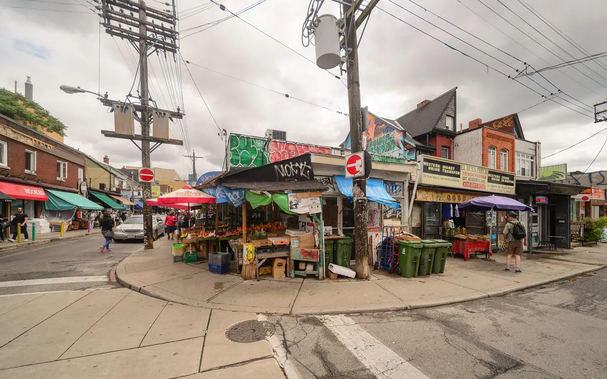 A fruit and vegetable stall under awnings at a Kensington Market corner, with graffiti-covered walls and shopfronts behind it