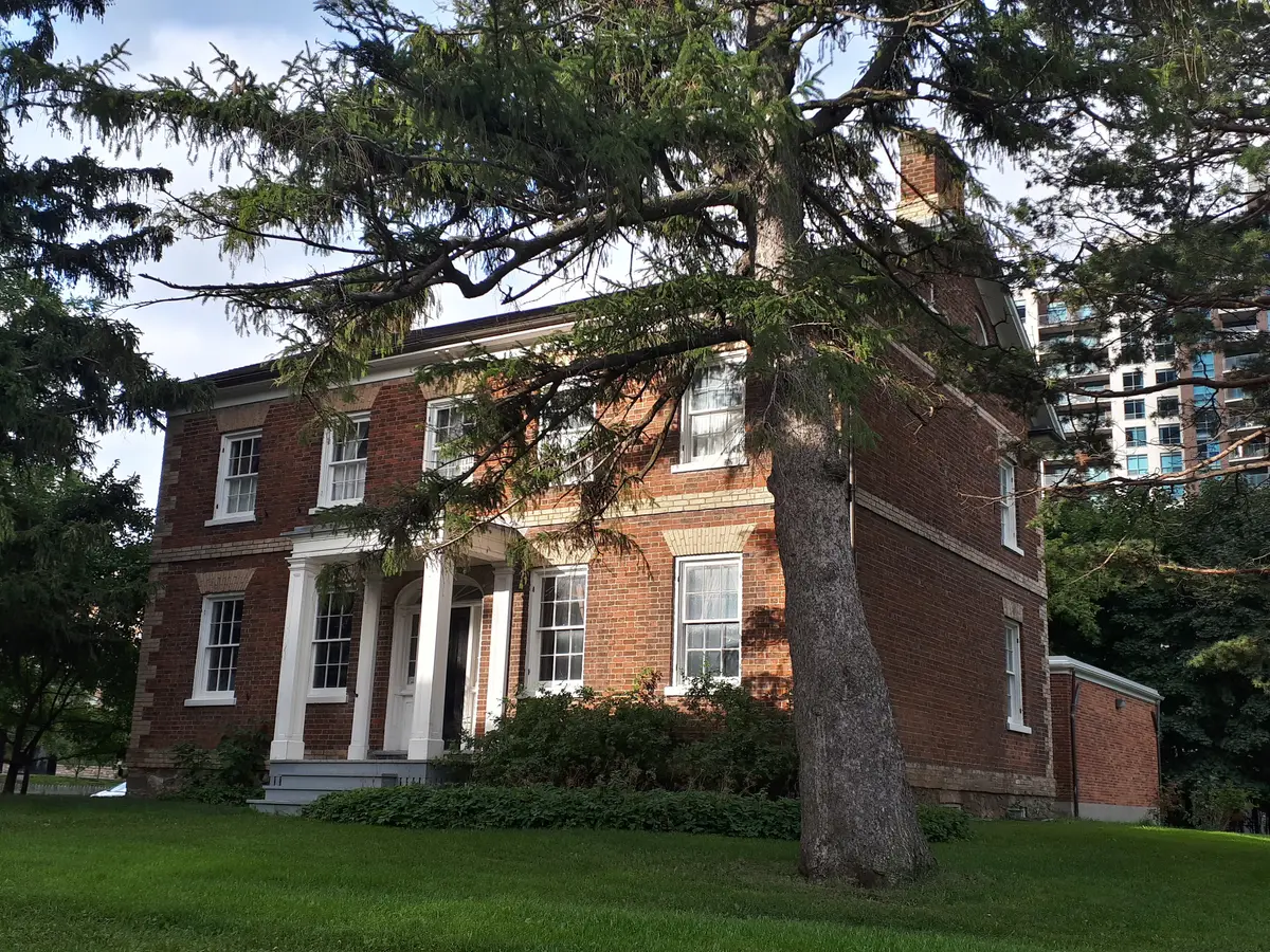 Red brick Gibson House with a white columned entrance, shaded by a large conifer, with high-rise apartment buildings behind it
