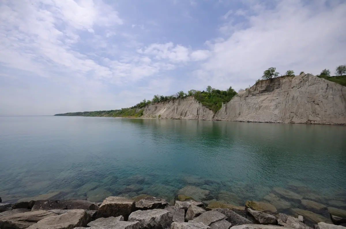 Pale clay cliffs of the Scarborough Bluffs rising above calm, clear lake water, with large rocks in the foreground and green trees along the top