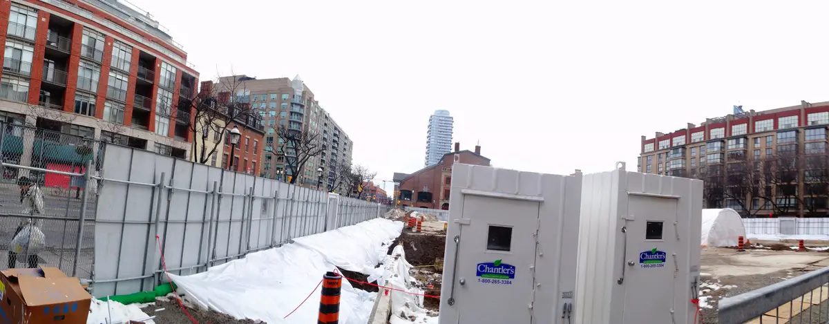 A long excavation trench lined with white sheeting, beside construction fencing, with brick and glass buildings beyond