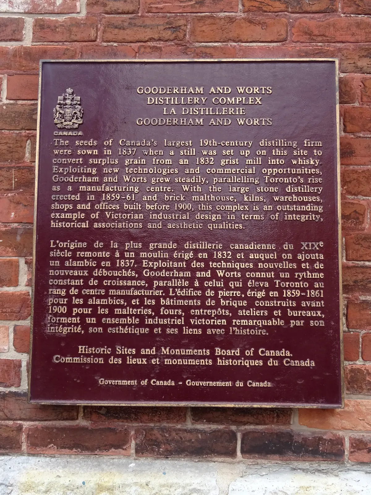 A maroon and gold Government of Canada heritage plaque on a red brick wall, in English and French