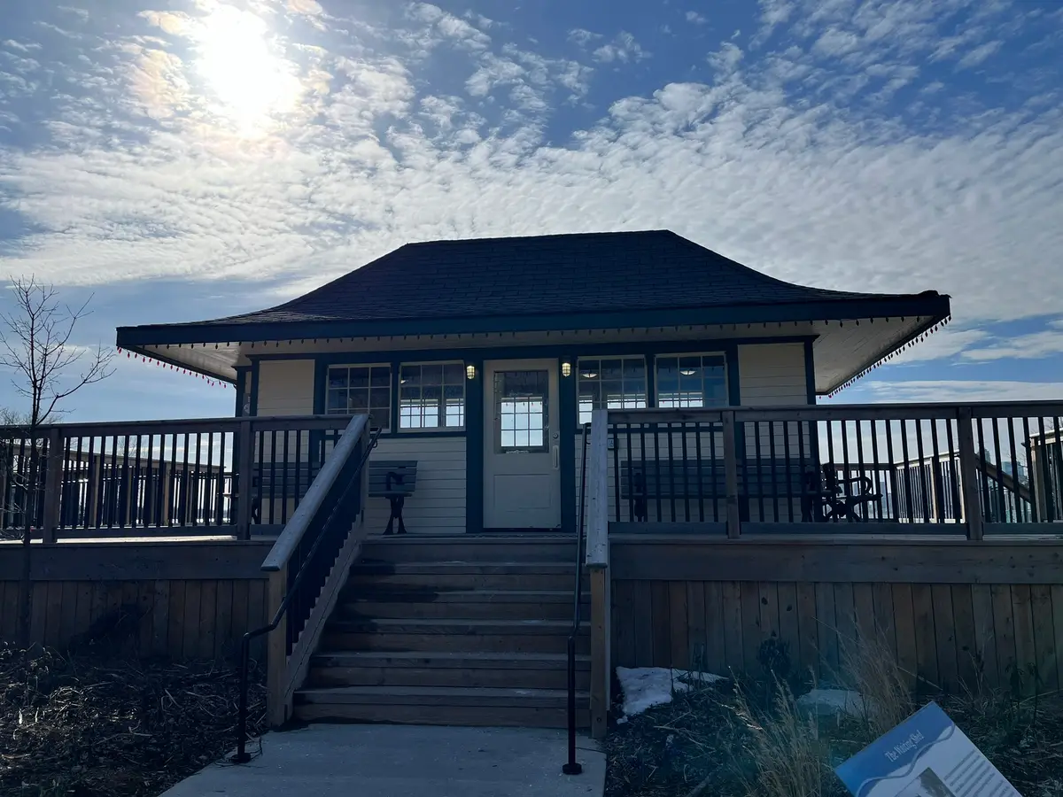 A small wooden waiting shed with a hip roof on a raised deck, with a sign reading The Waiting Shed in front, under a blue sky
