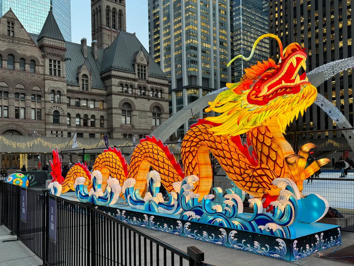 A glowing orange and gold lantern dragon on blue waves beside the rink at Nathan Phillips Square, with Old City Hall behind