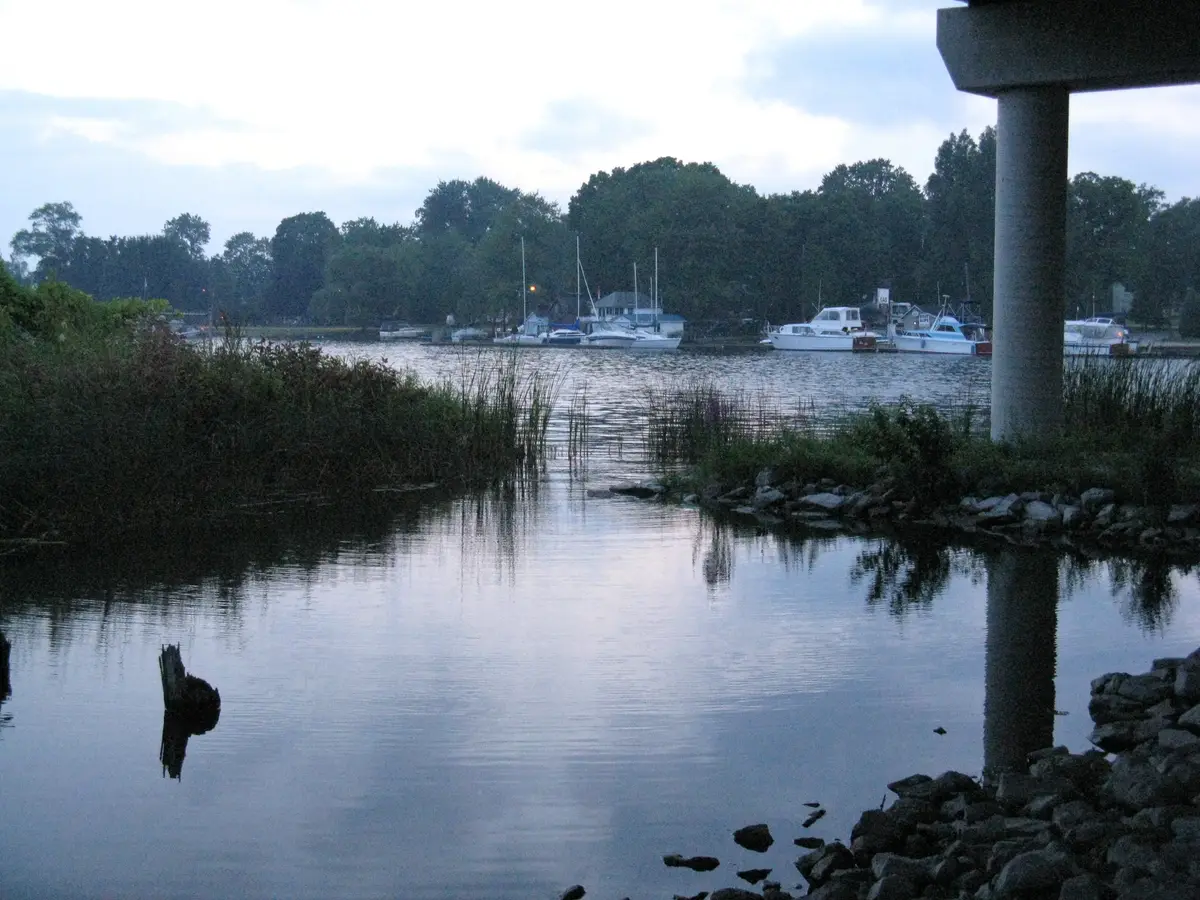 A calm channel at dusk with reeds, rocks and moored boats, and a bridge pier at the right of the frame