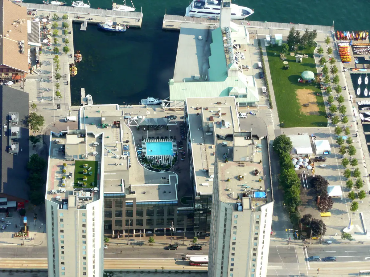 Aerial view of a waterfront with a marina, a rooftop pool and sun terraces between two high-rise towers, a lawn beside a row of kayaks, and a streetcar on the road below