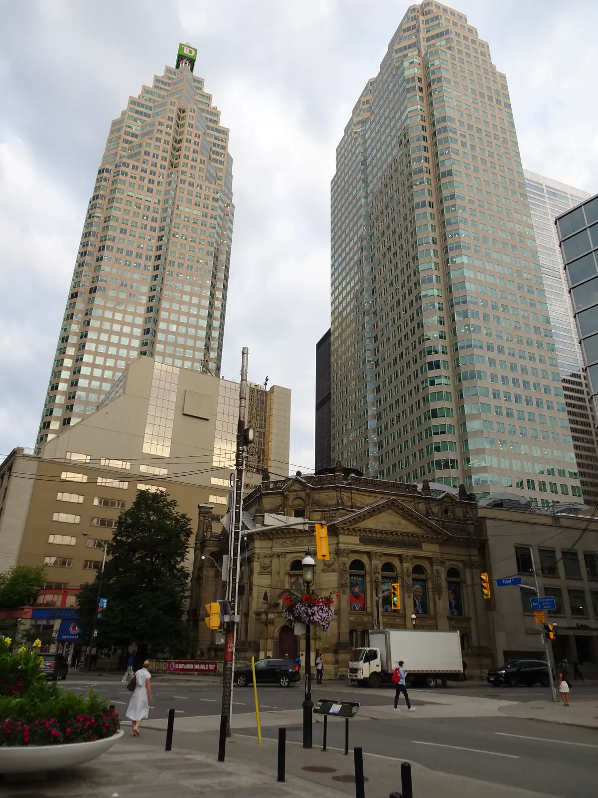 City street corner with a sandstone building and two glass towers behind it, and a small plaque on a post at the curb in the foreground