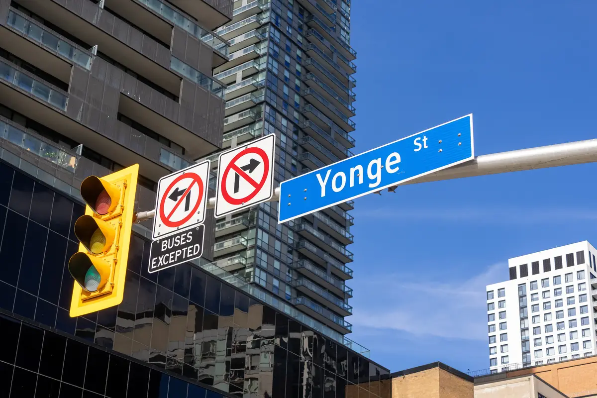 A blue Yonge Street sign on a mast arm beside a traffic signal and turn-restriction signs, with glass towers behind
