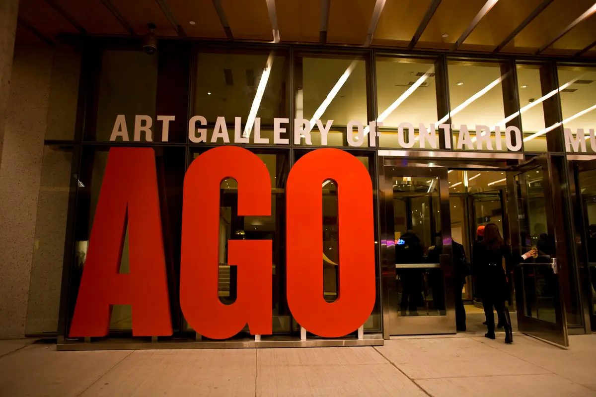 Large red AGO letters in front of the glass entrance of the Art Gallery of Ontario, with a visitor at the door.