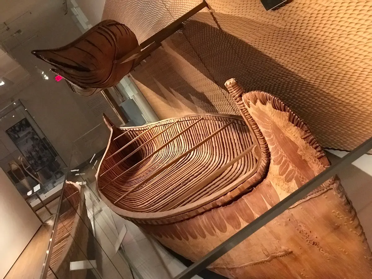 A birchbark canoe in a glass display case, with woven and textured panels behind it.
