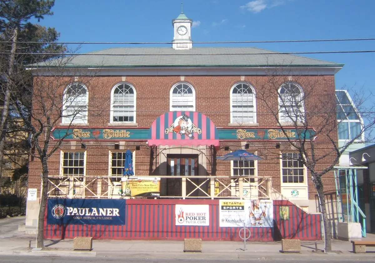 A two-storey red brick building with arched windows and a small clock cupola on Dundas Street West, with a patio fence at street level