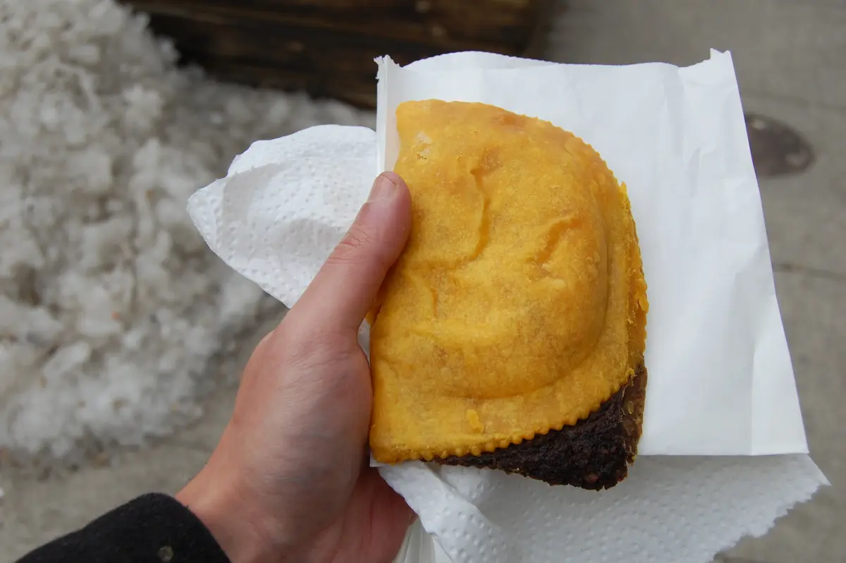 A golden Jamaican patty held in one hand on a paper wrapper, with snow on the ground behind