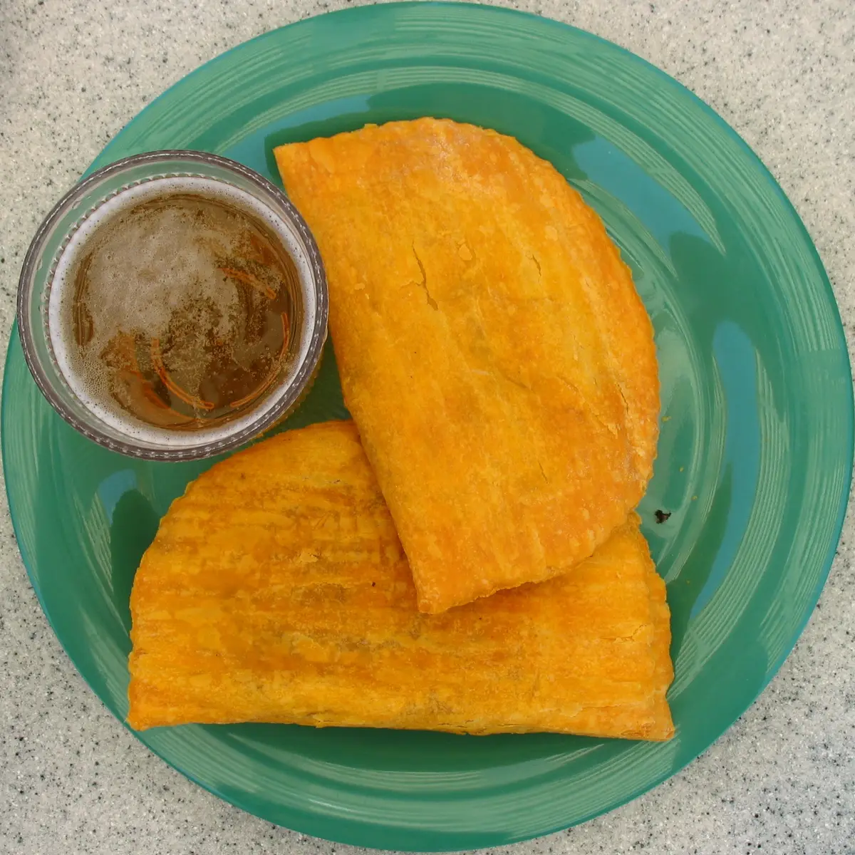 Two golden Jamaican patties on a green plate beside a glass of drink