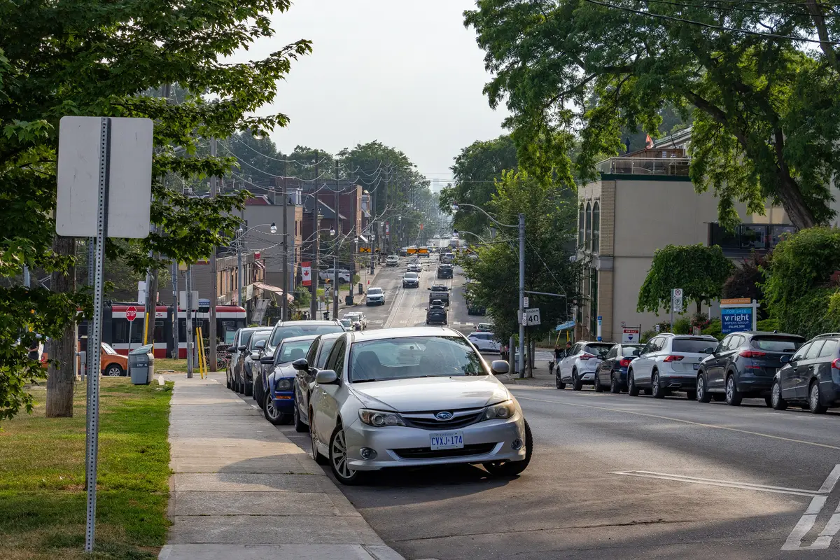 Queen Street East in the Beaches, with parked cars along the curb, a red streetcar and tree-lined storefronts under a hazy summer sky