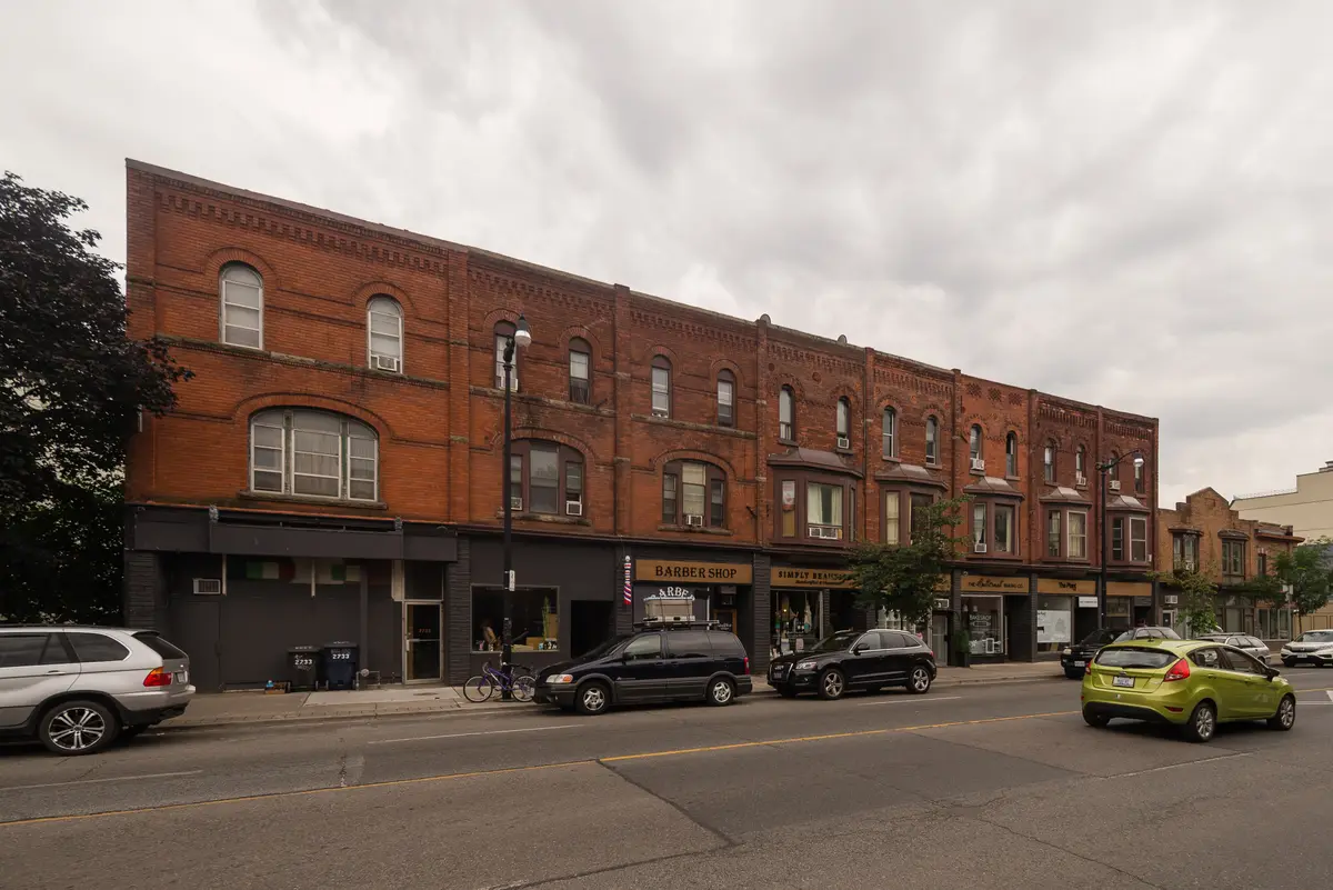 Three-storey red-brick shopfronts on Dundas Street West, with a barber shop sign and parked cars along the street
