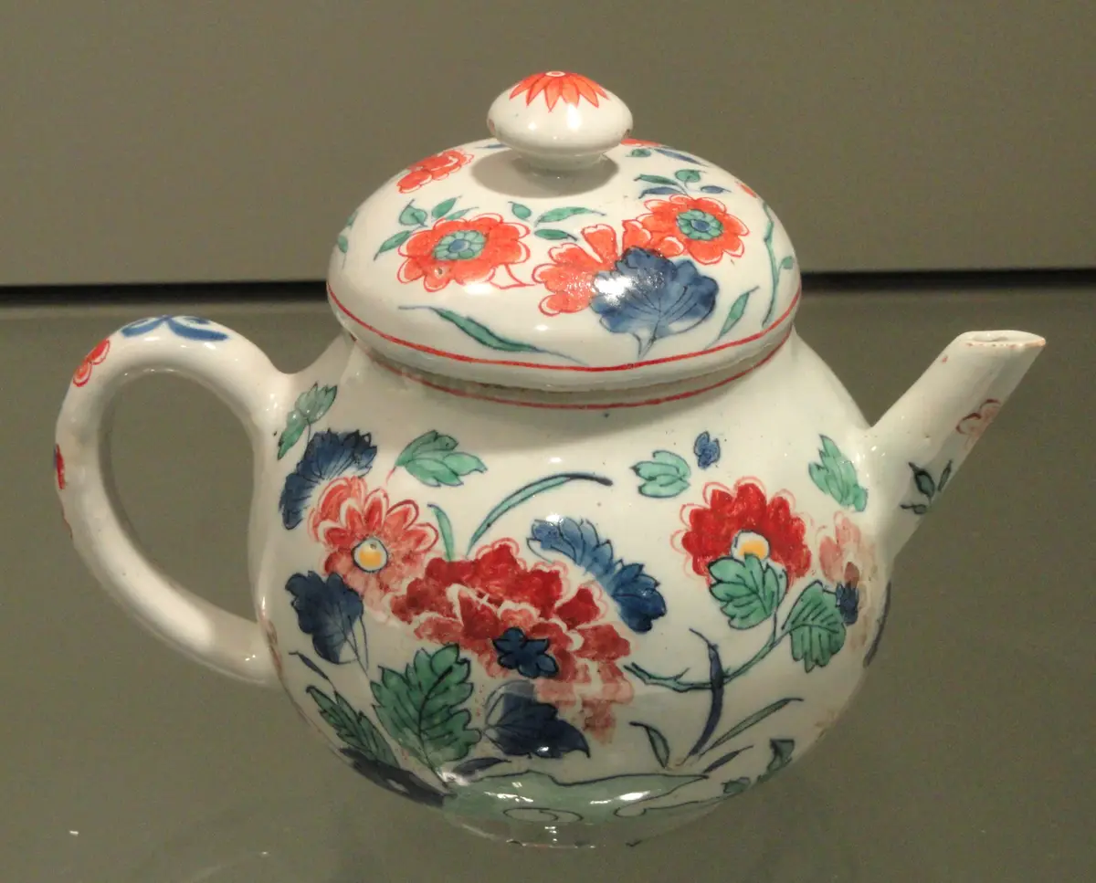 A white teapot with a domed lid and a short spout, painted with red, blue and green flowers and leaves.