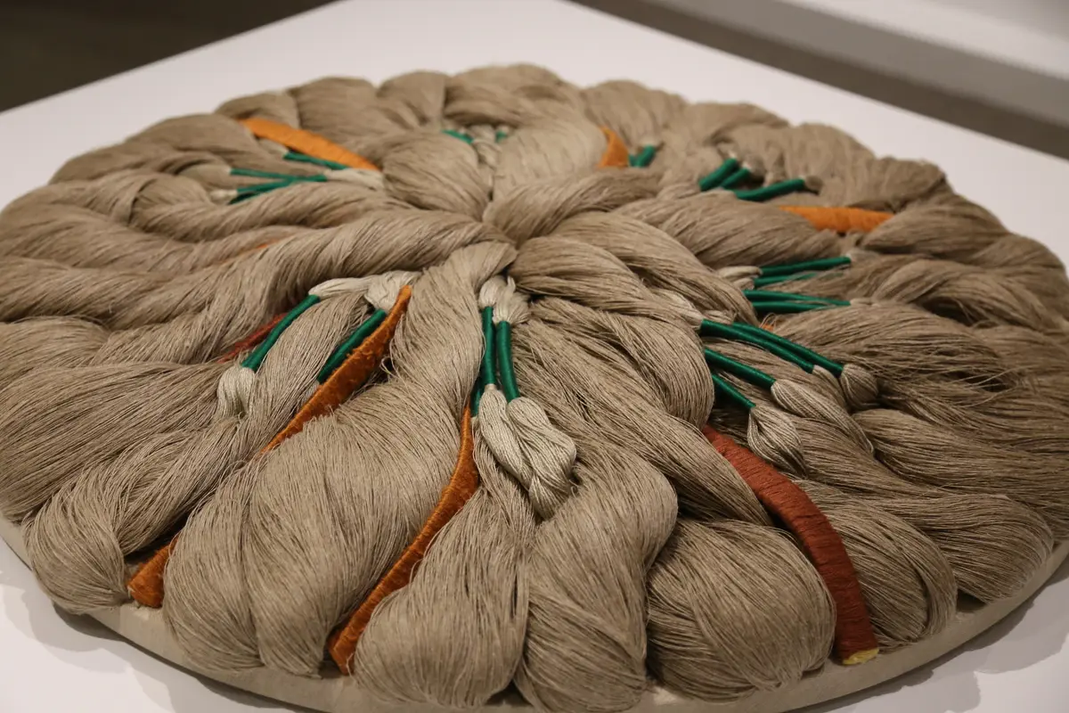 A round textile work of bundled undyed fibre with orange, green and rust cords, laid on a white display surface, from a past Sheila Hicks exhibition at the Textile Museum of Canada photographed in 2016.