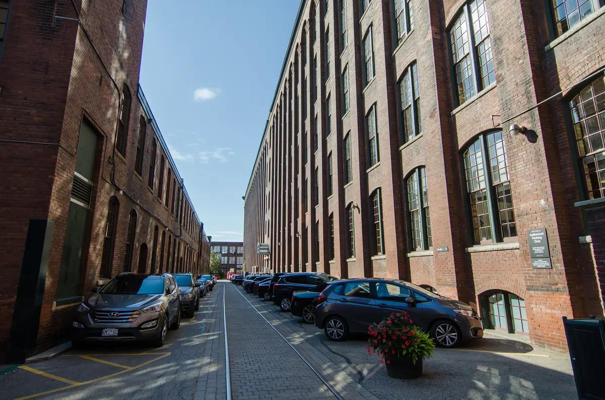 A paved laneway running between tall brick factory buildings with parked cars along one side
