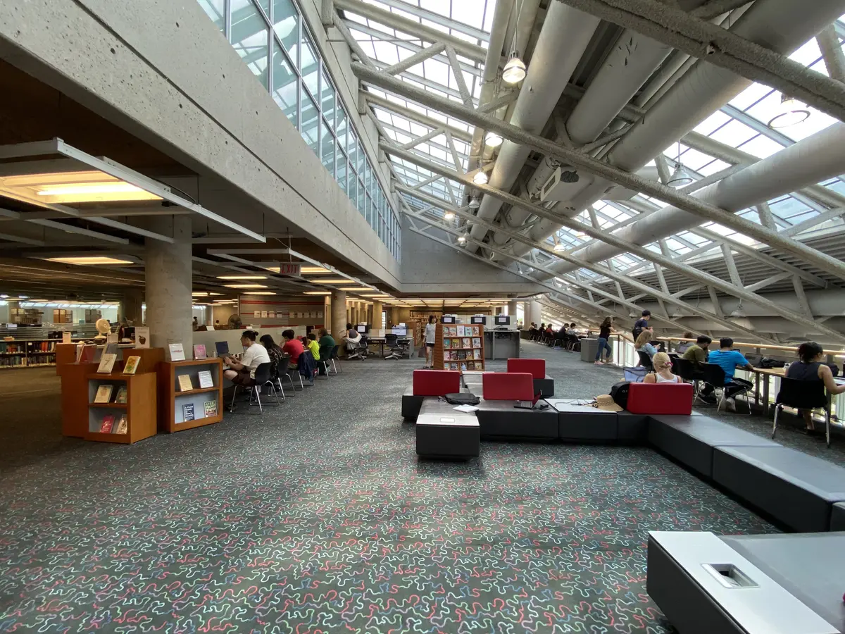 Reading area in the Fairview branch with people working at tables, red and black seating in the foreground, and exposed ceiling trusses with skylights overhead.