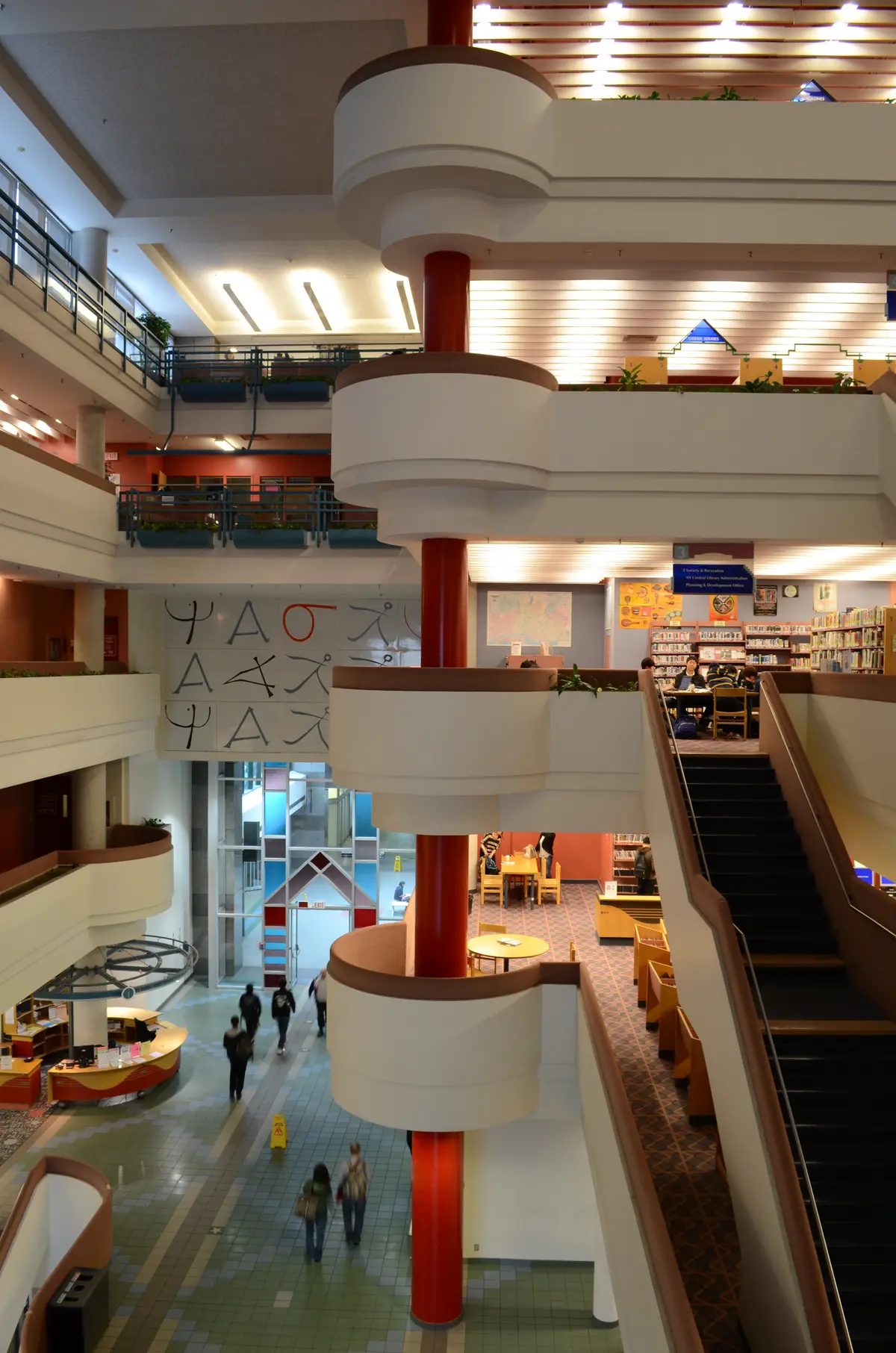 Atrium at North York Central Library, with curved white balconies around a red central column, a staircase on the right and people walking across the ground floor.