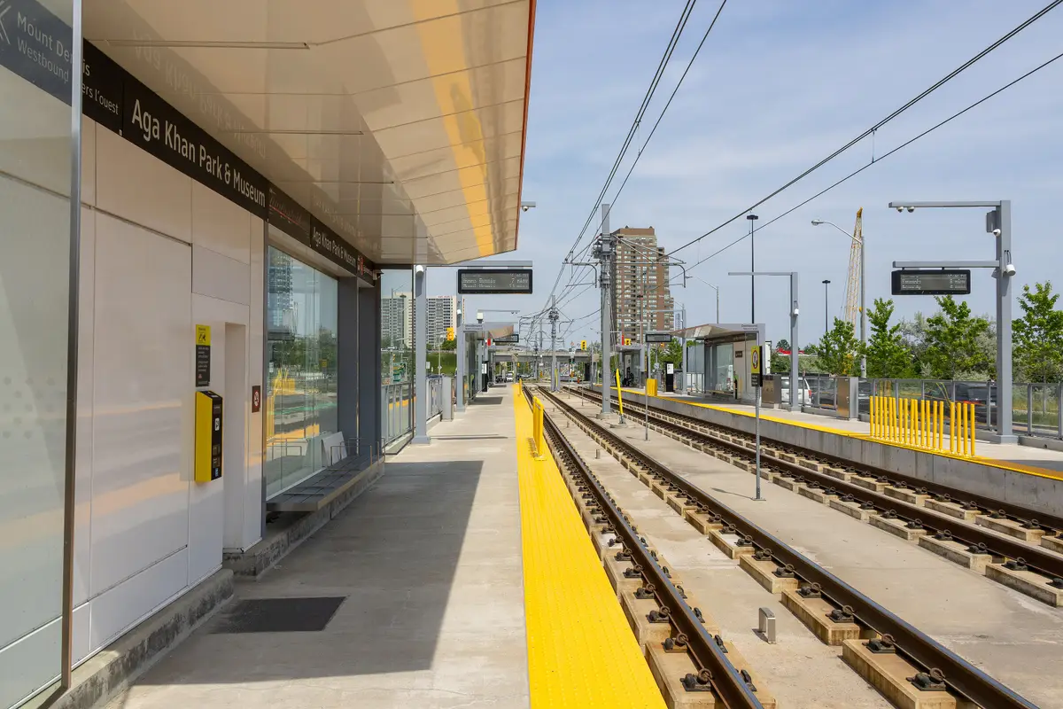 The westbound platform at Aga Khan Park & Museum station, with the station name on the shelter wall and the tracks running ahead under a blue sky.