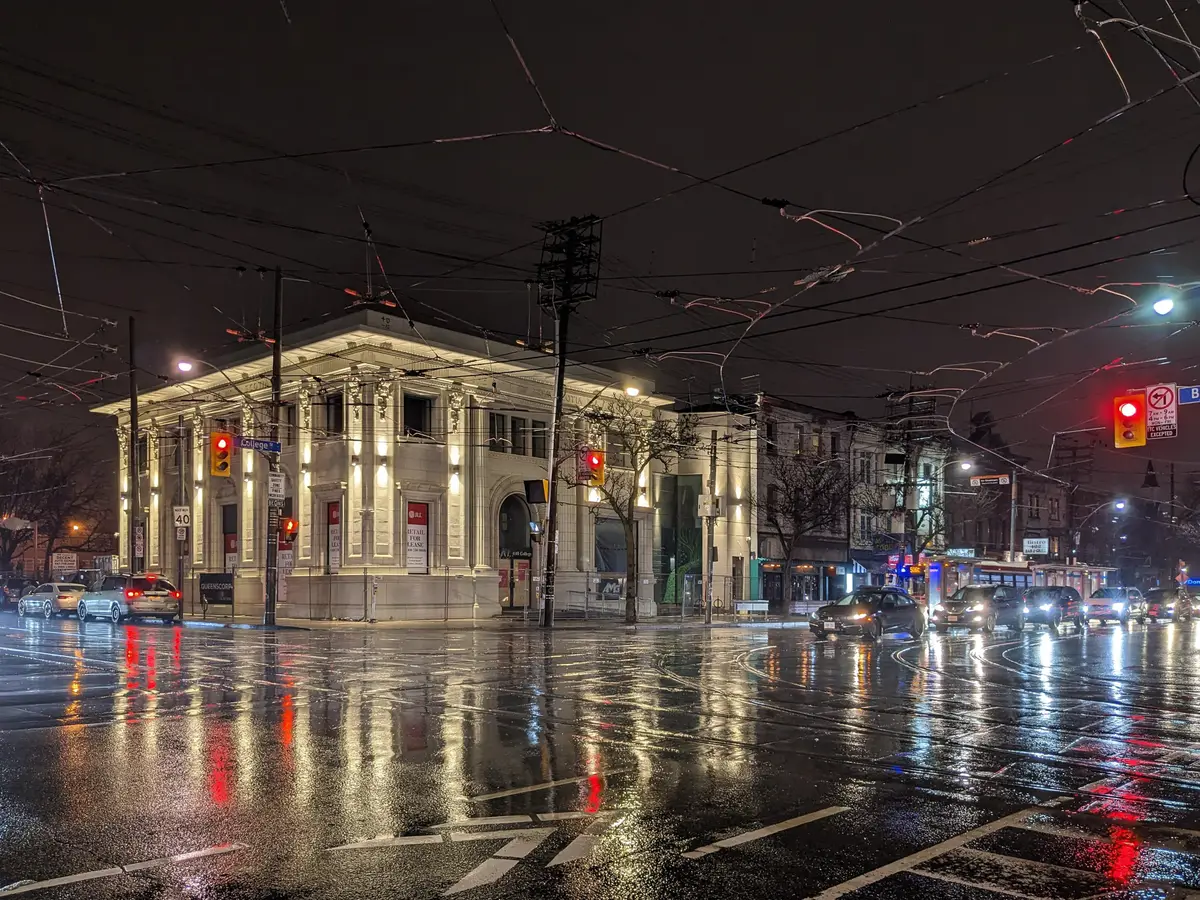 A lit stone corner building and wet pavement at College Street and Bathurst Street on a rainy night